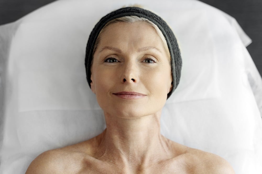 	
A middle-aged woman with bare shoulders and a headband lies on a treatment bed, looking up with a relaxed expression, perhaps reflecting on the potential facelift benefits and risks involved in her rejuvenation journey.