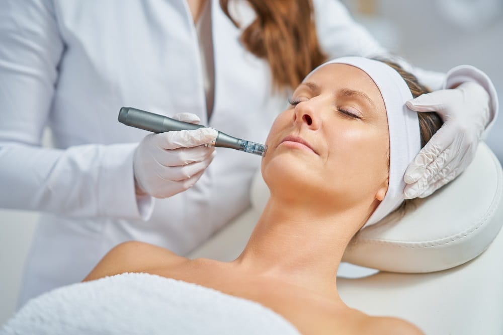 A person receives a facial treatment with a blue-light therapy device, while a professional wearing gloves prepares for microneedling for wrinkles.