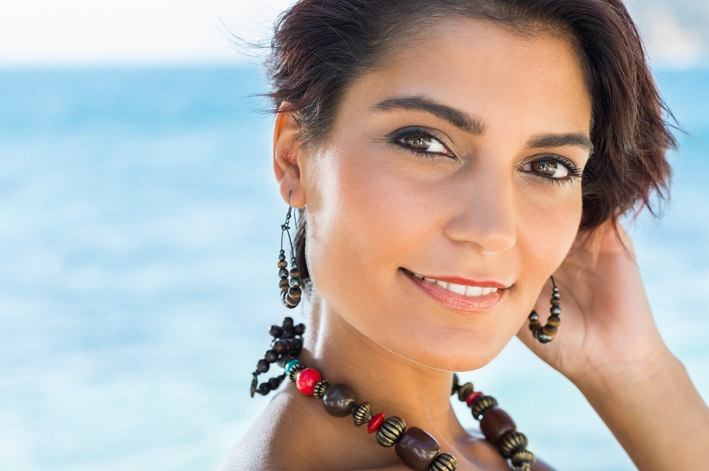 A woman with short dark hair wearing beaded jewelry smiles at the camera with the ocean in the background.