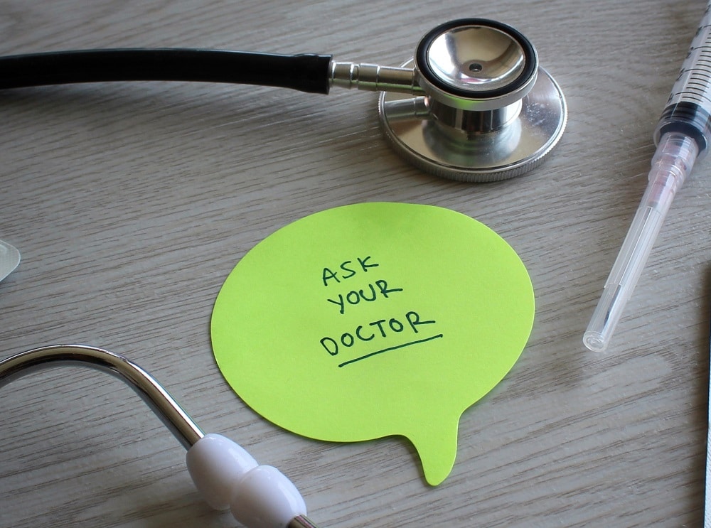 A stethoscope, syringe, and a green speech bubble note with "ASK YOUR DOCTOR" remind you to consider important questions to ask surgeons, all arranged neatly on a wooden surface.